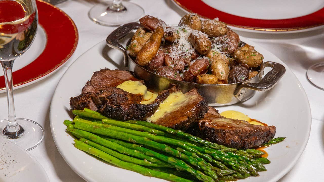 A plated meal with steak topped with butter, roasted potatoes in a dish, and asparagus, accompanied by glasses of wine.