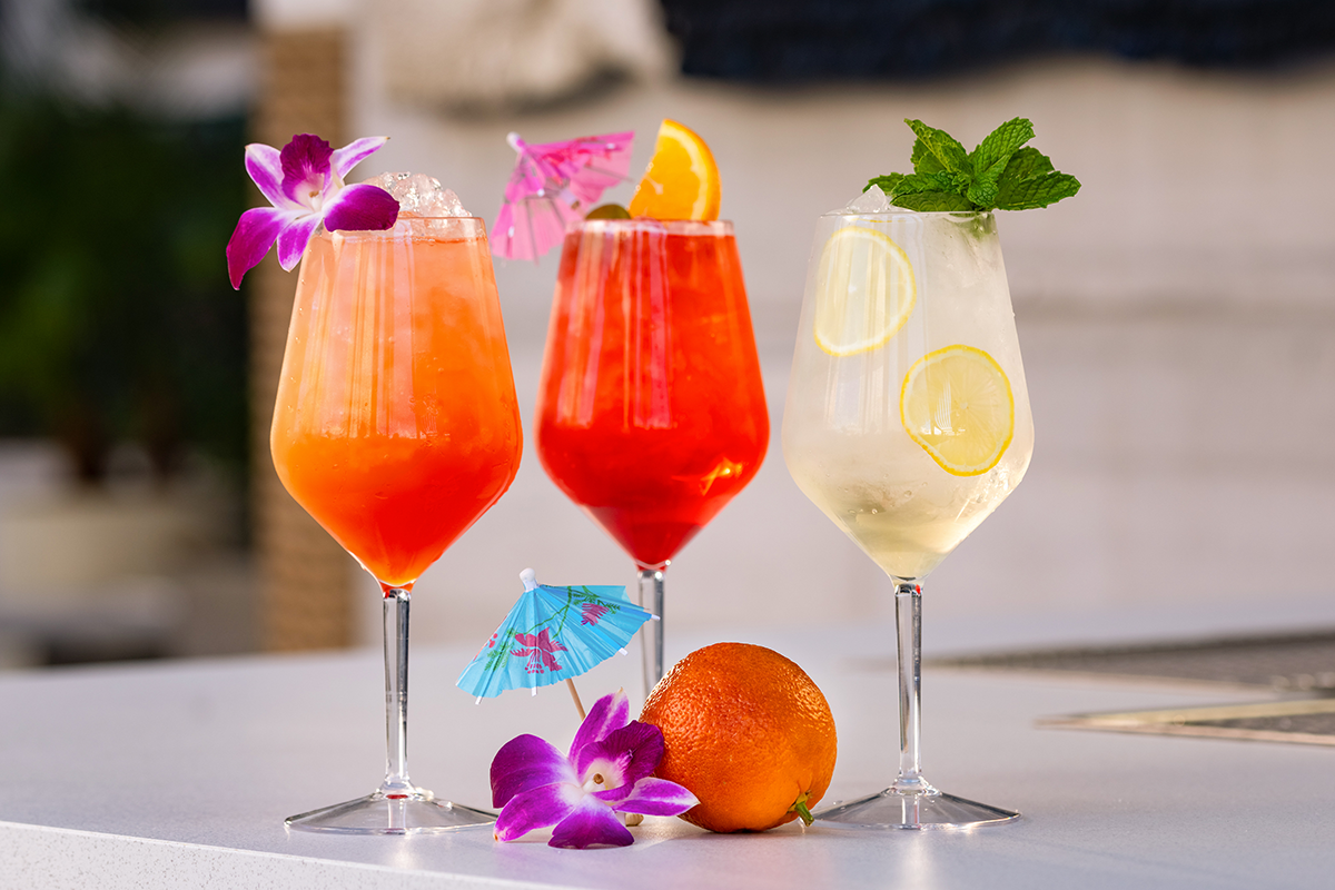 Three colorful cocktails in tall glasses, garnished with flowers, umbrellas, orange slices, and mint, set on a white surface.