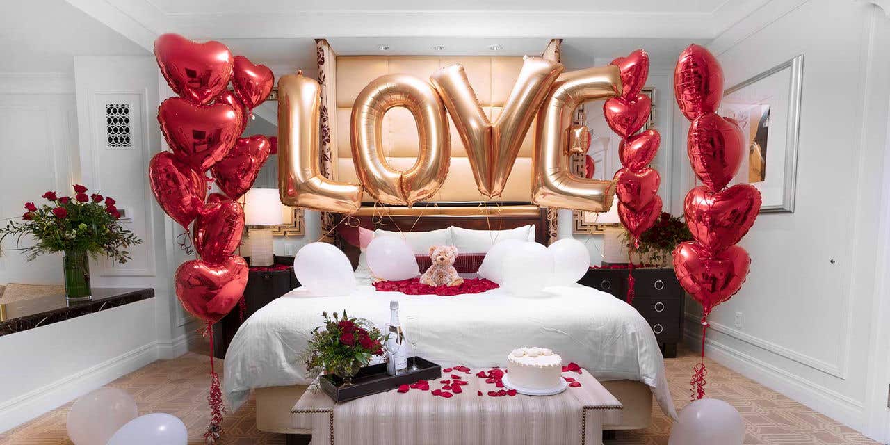 A hotel bedroom with flowers, heart shaped balloons, and mylar balloons spelling “love” surrounding the bed.
