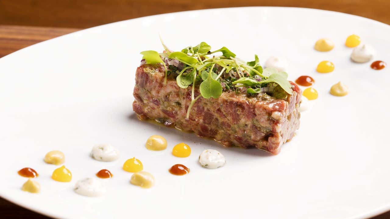 A gourmet dish featuring steak tartare topped with microgreens, surrounded by colorful sauce dollops on a white plate.