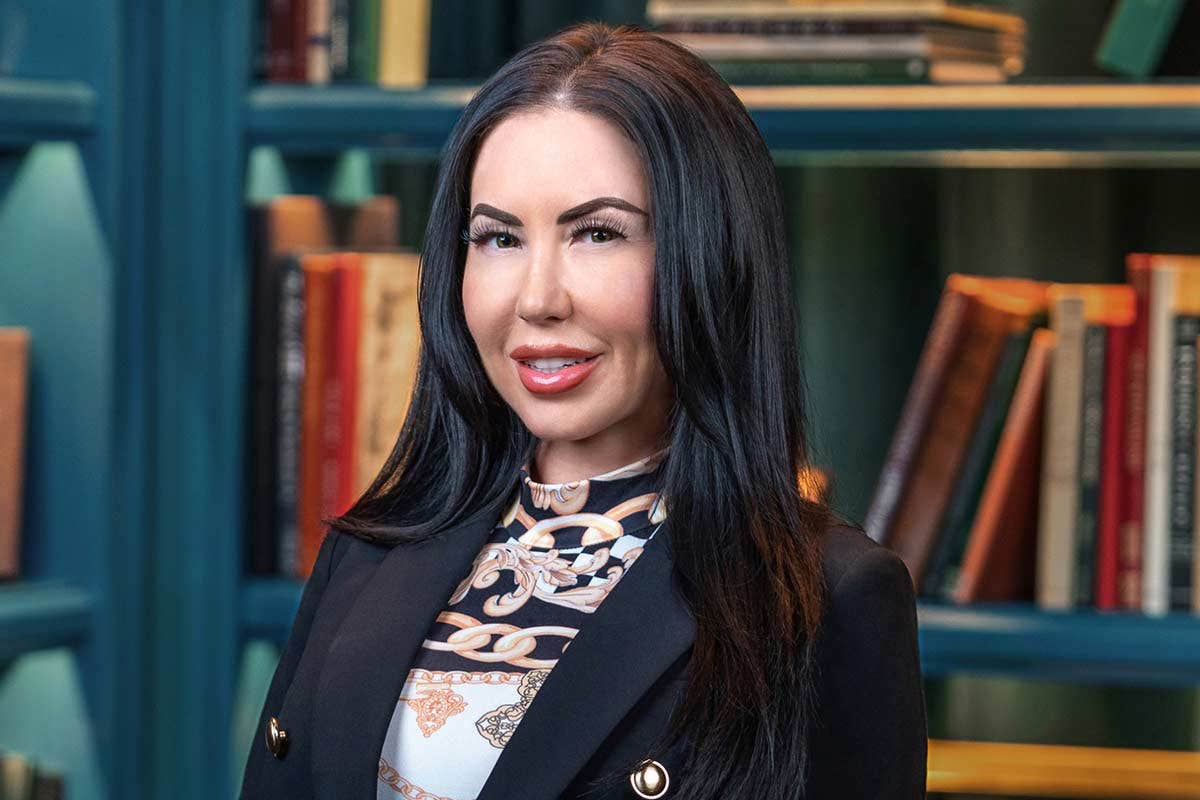 A person with long dark hair wearing a patterned blouse and blazer, standing in front of a bookshelf.