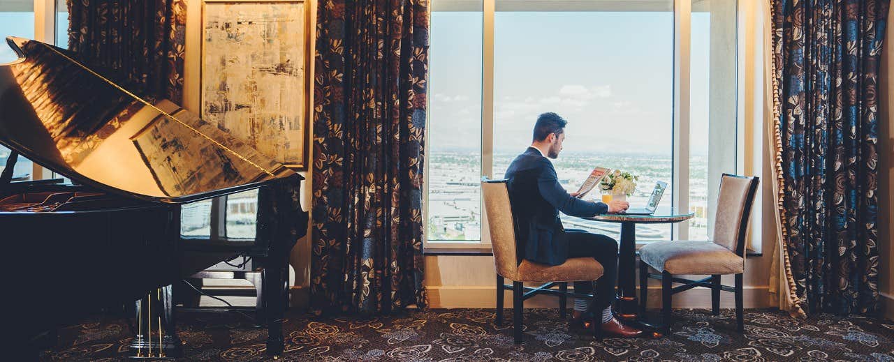 A man reads a book while seated at a round table in a high-rise room with a view, next to a grand piano.