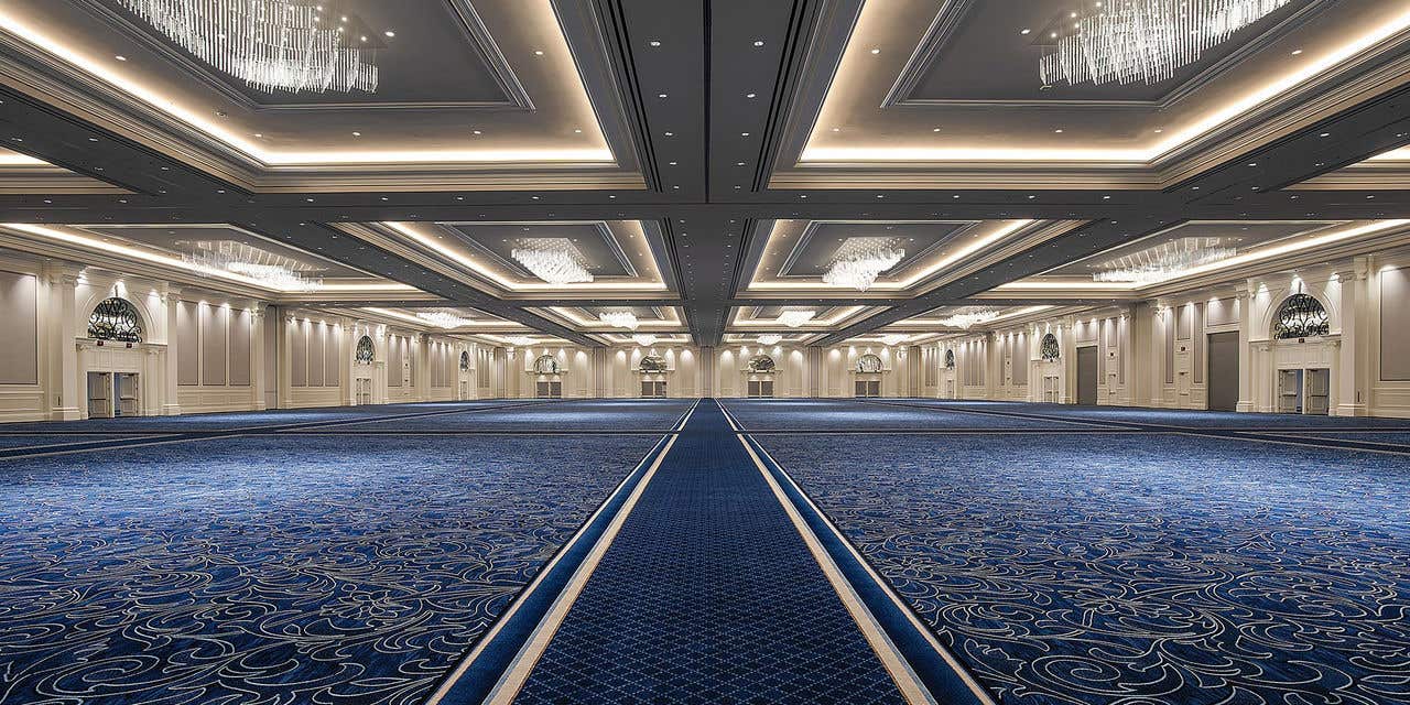 Large empty ballroom with white walls, blue carpet and chandeliers hanging from the ceiling.