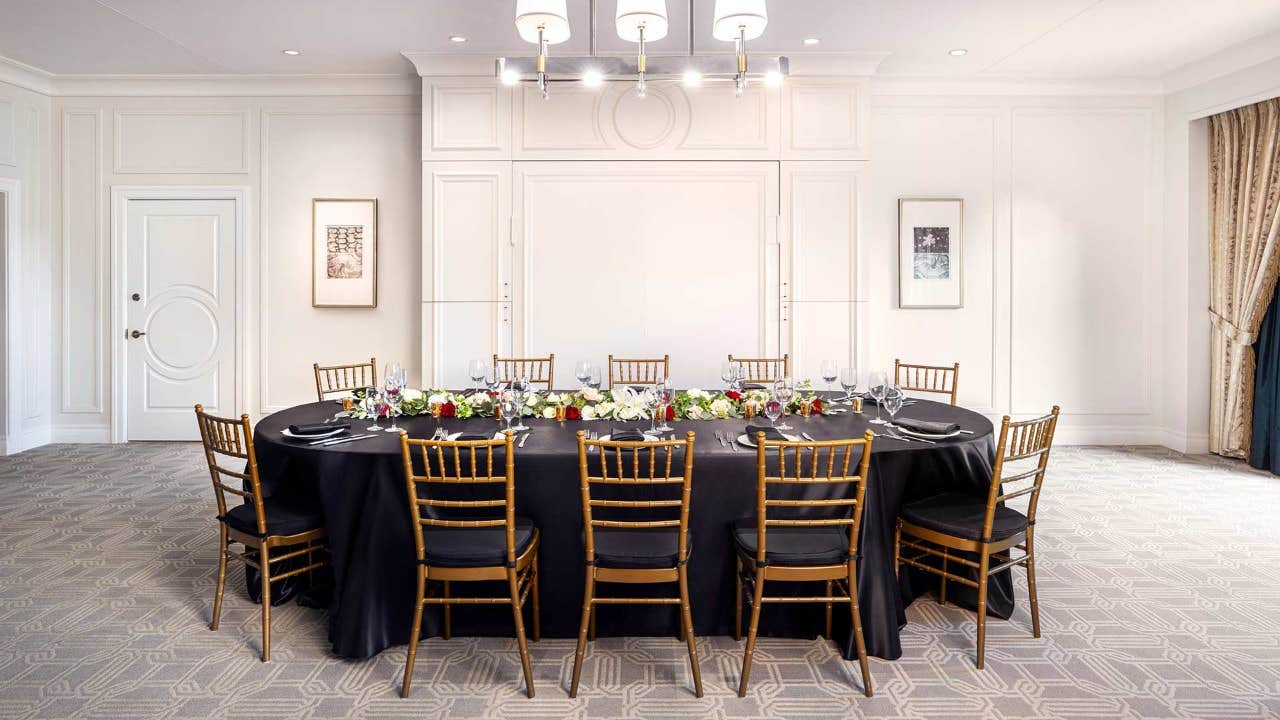 A round dining table with eight chairs, set with black tablecloth, elegant tableware, and floral centerpieces in a bright room.