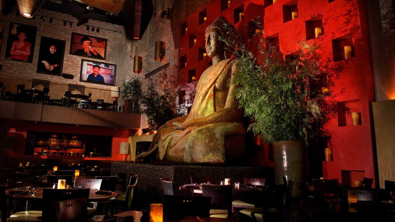 A restaurant interior with a large Buddha statue, ambient lighting, potted plants, and framed portraits on the walls.