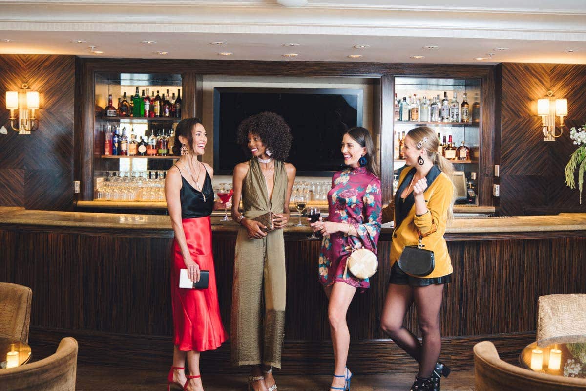Four women stand at a bar with drinks while talking and laughing.