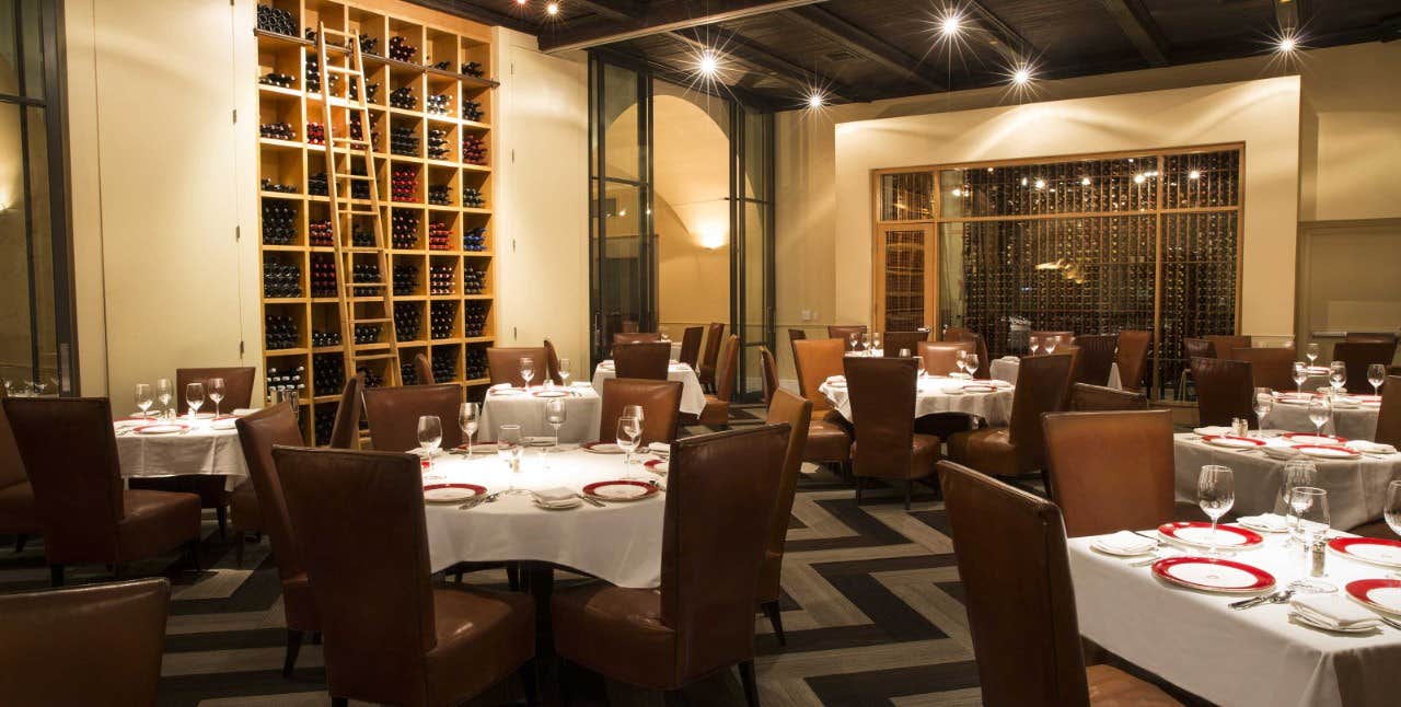 Elegant restaurant interior with leather chairs, round tables with white cloths, and shelves filled with wine bottles on walls.