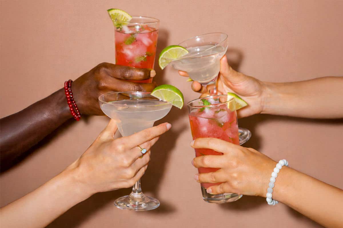 Four hands clink assorted cocktails against a plain peach-colored background, including two clear margarita-style drinks and two tall pink beverages with visible ice and mint. Each glass is garnished with a lime wedge, and some hands wear simple bracelets and rings.