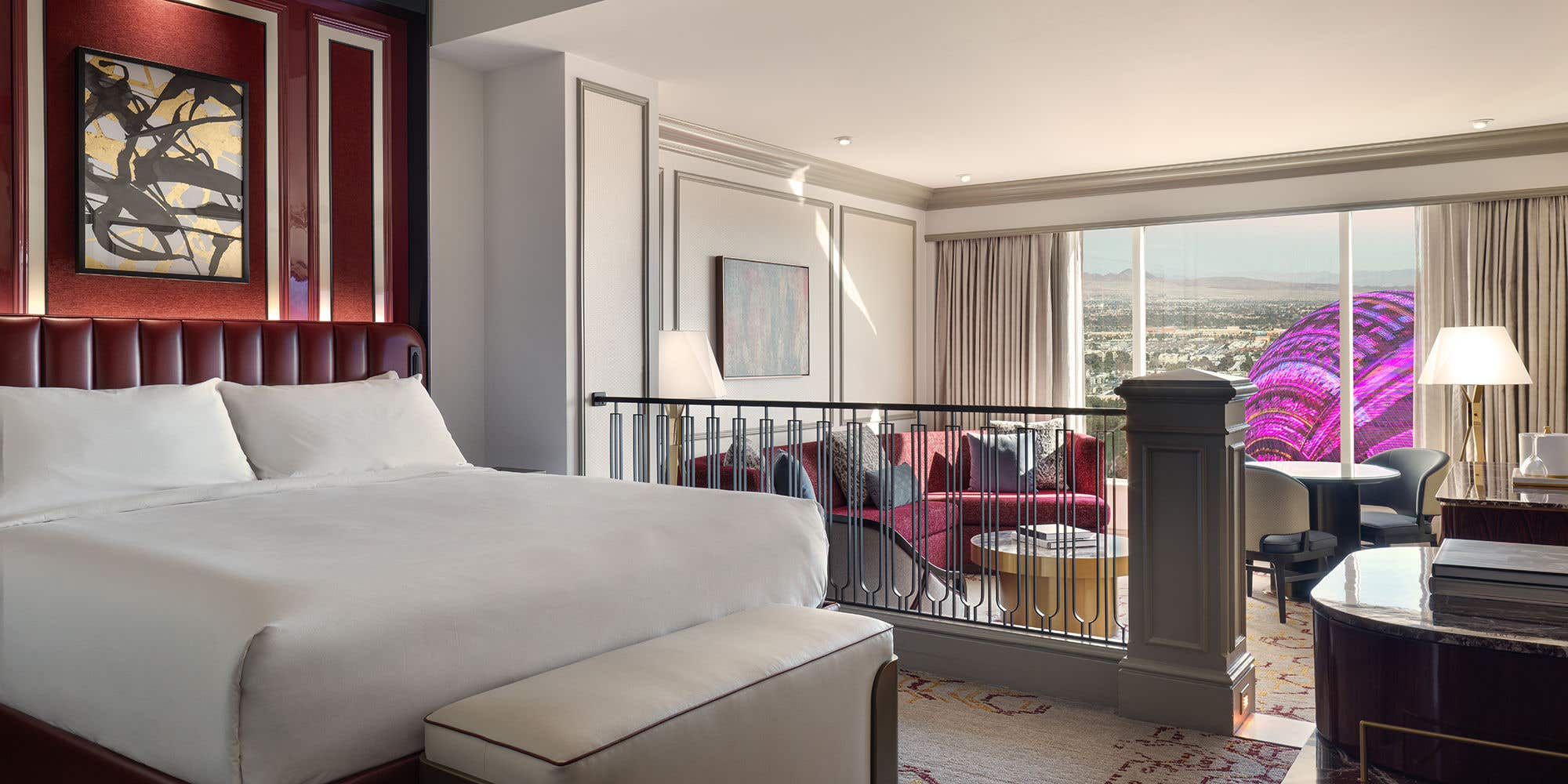 Remodeled Luxury King Suite bedroom with a bench seat at the foot of the bed. In the background is a sunken living room with an armchair, couch, coffee table, dining table with chairs, lamps, and a daytime view of Sphere at The Venetian outside the window.