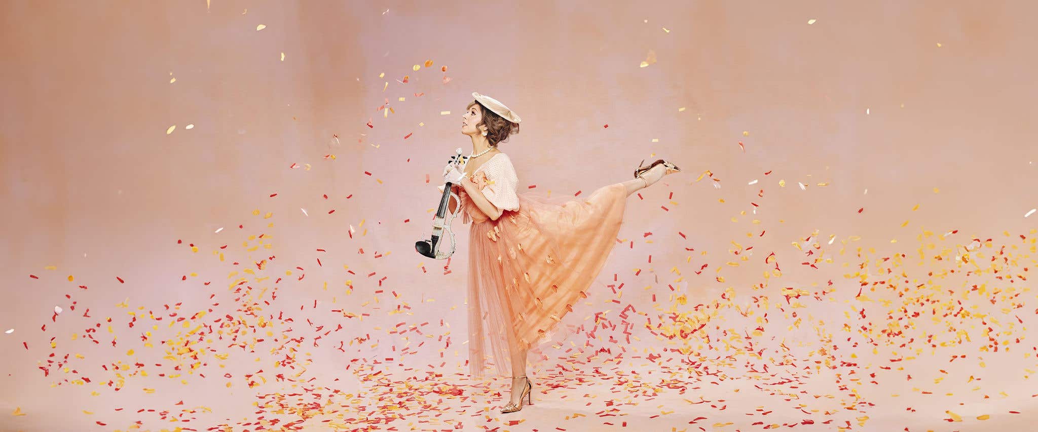 Lindsey Stirling posing with an electric violin standing on one leg whilst against a light orange backdrop with confetti