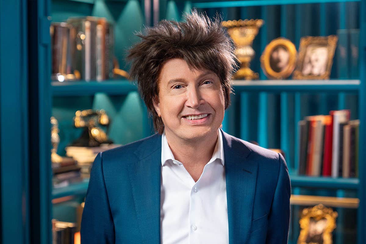 A person with a smile, wearing a blue suit, stands in front of a bookshelf filled with books and decorative items.