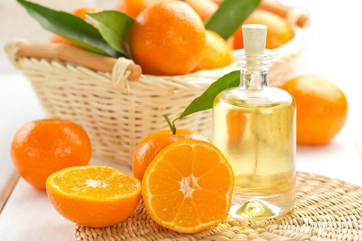 A bottle of citrus oil next to fresh oranges, some whole and one halved, on a woven mat with a basket of oranges in the background.