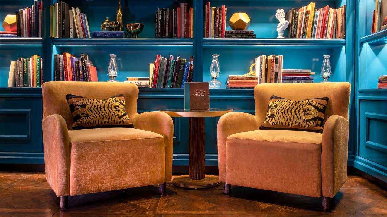 Two orange armchairs with tiger cushions face a small table, set against a blue bookshelf filled with colorful books.