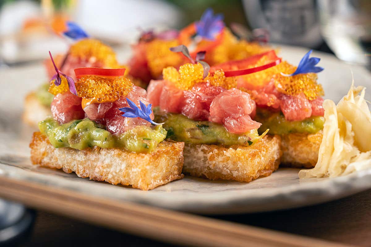 A close-up of crispy rice squares topped with avocado mash, spicy tuna, golden tobiko, and edible purple flowers, served with pickled ginger on a white ceramic plate. 