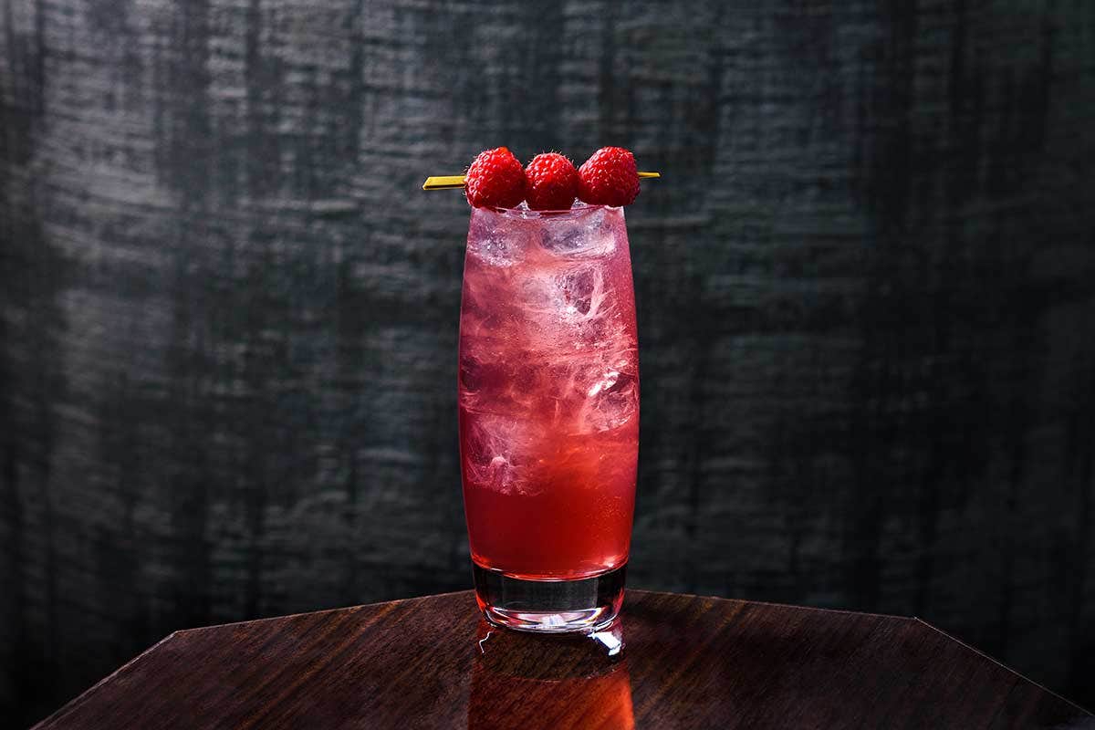 A tall glass of red cocktail on a wooden table, garnished with three raspberries on a skewer.