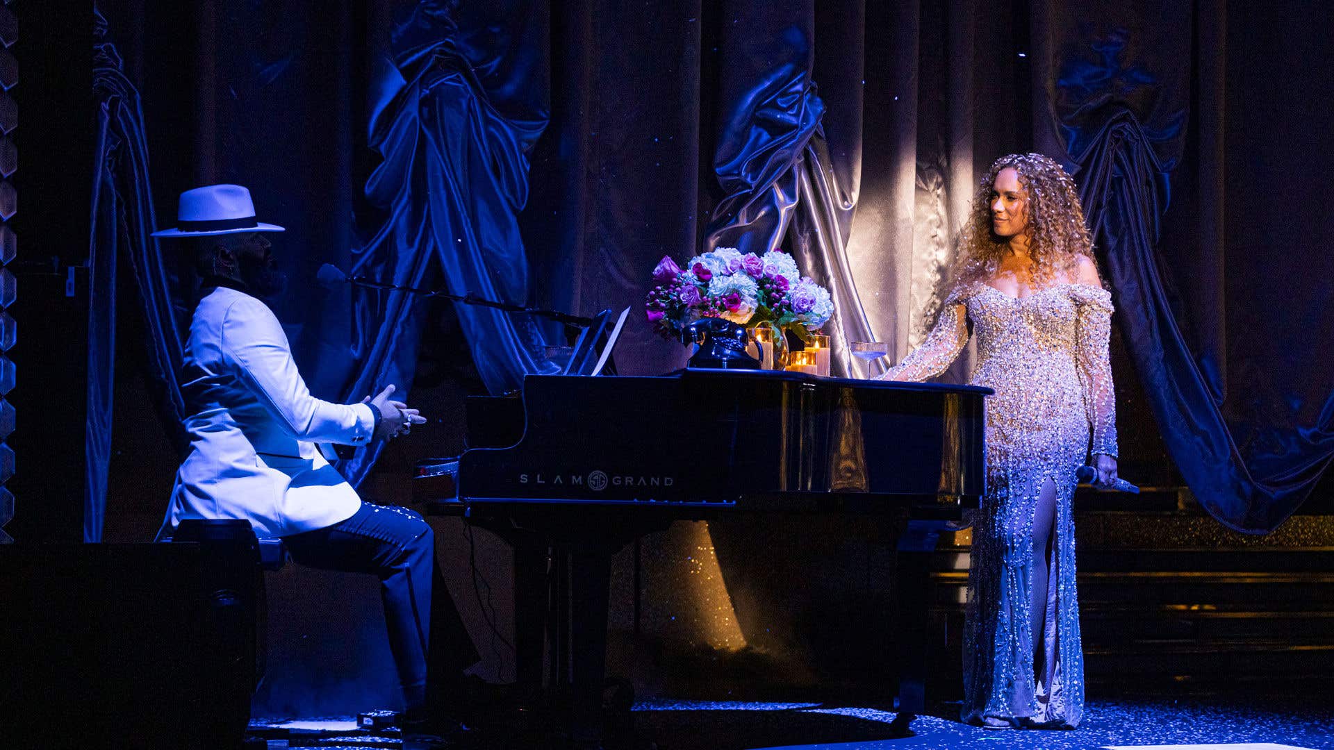 Leona Lewis during her Christmas show in Las Vegas wearing a silver sequin dress and leaning against a black grand piano with a pianist in a white suit.