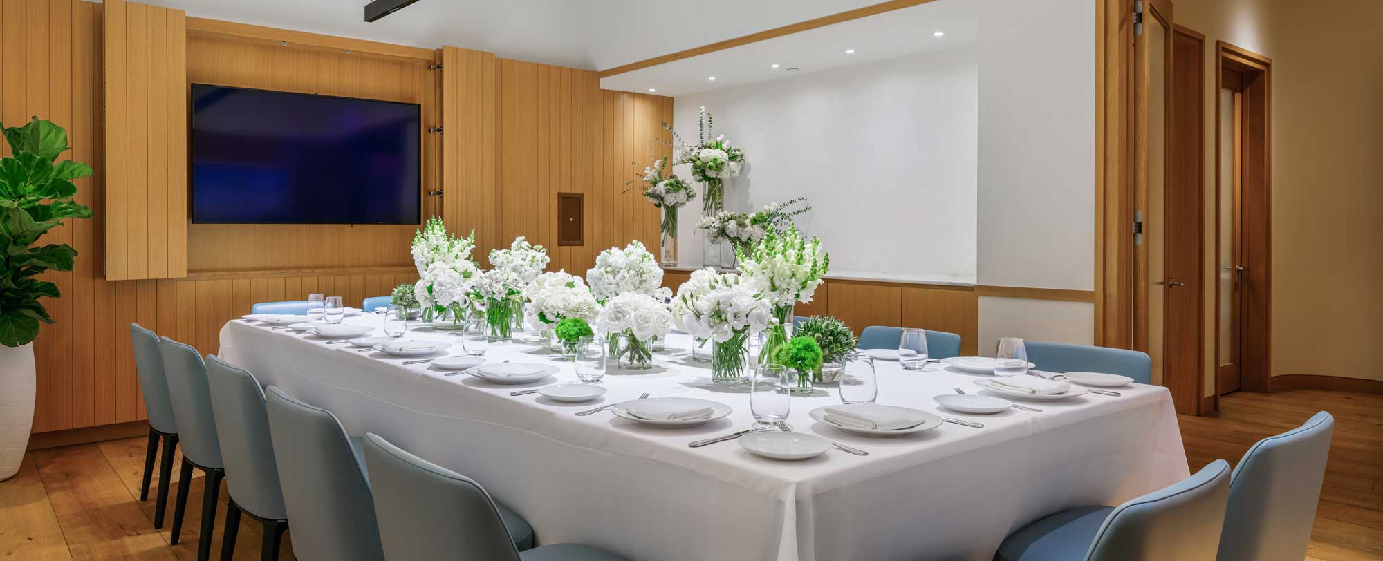 A modern dining room with a long table set for a meal, adorned with white flowers, plates, and a large TV on the wall.