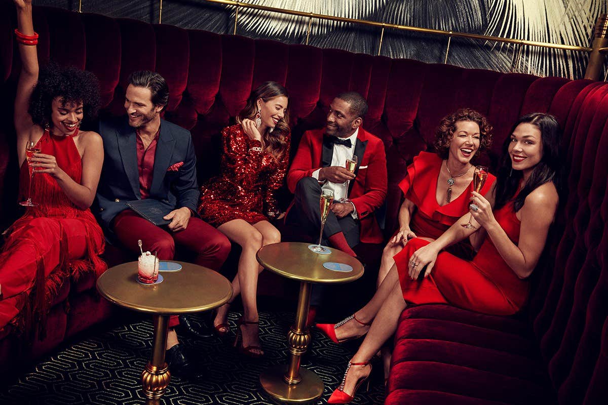 A group of elegantly dressed people in red and black enjoy drinks in a luxurious lounge with plush, red seating.