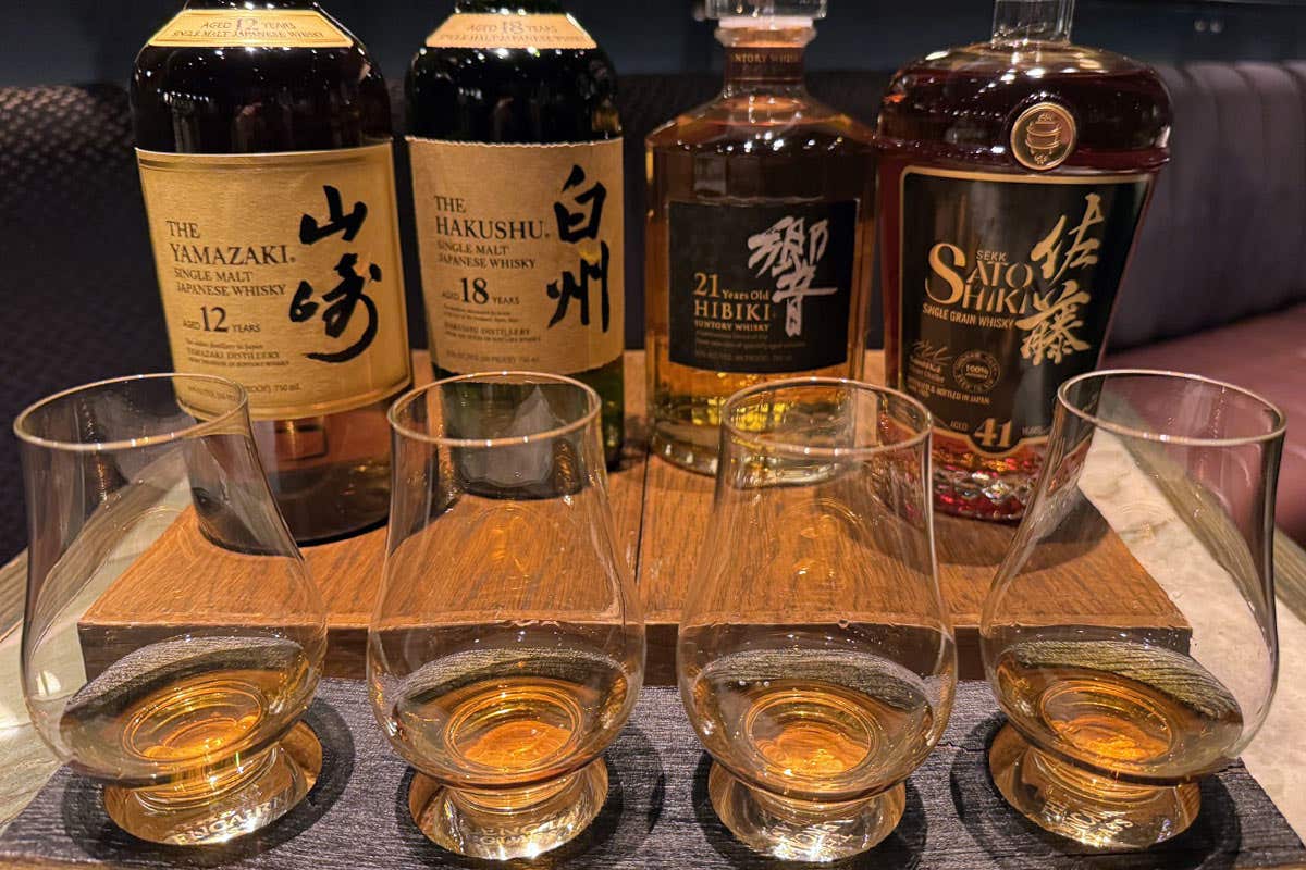 A whiskey tasting flight with four glasses of whiskey and their corresponding bottles, neatly arranged on a dark mat in a bar.