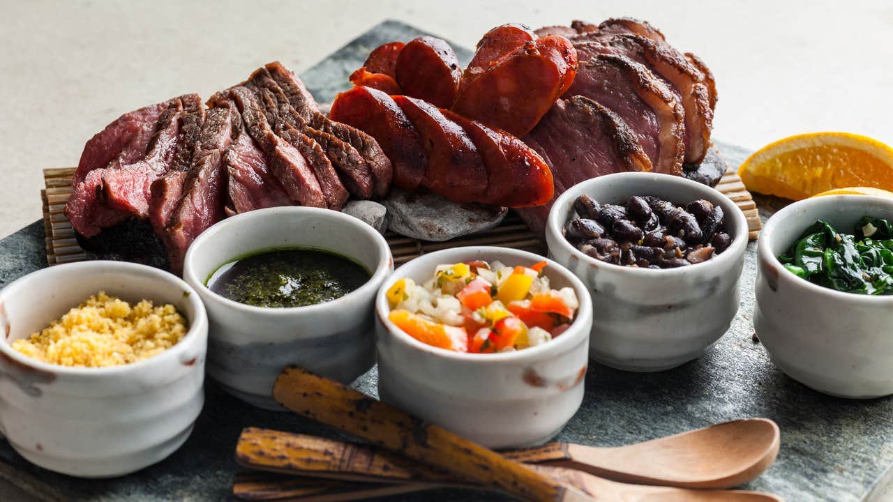 A platter of grilled meats with sides of sauce, beans, greens, salsa, farofa, and a wedge of orange, served on a stone board.