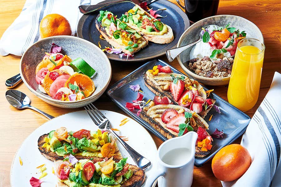 A table set with colorful avocado toasts, fruit bowls, a yogurt parfait, fresh orange juice, and oranges.