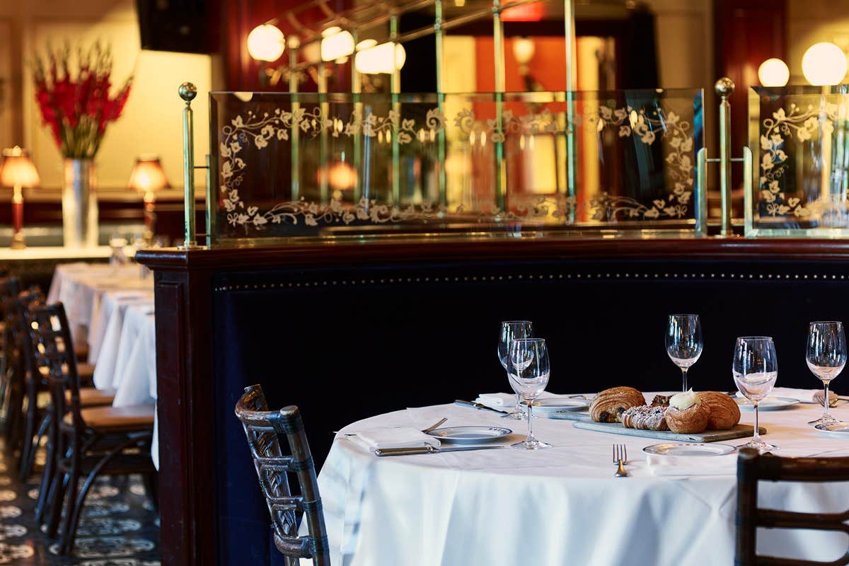 Elegant restaurant with a set table, empty wine glasses, and bread rolls. Warm lighting and a decorative divider in the background.