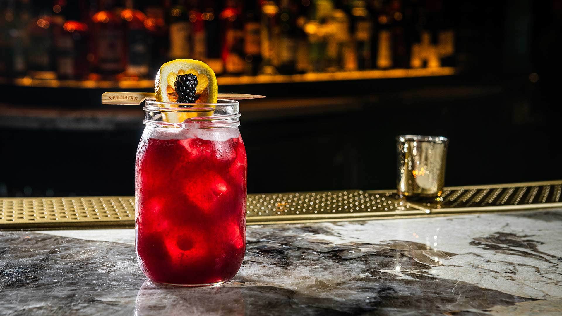 A dark red cocktail with ice, garnished with a lemon slice and blackberry, on a marble bar counter.