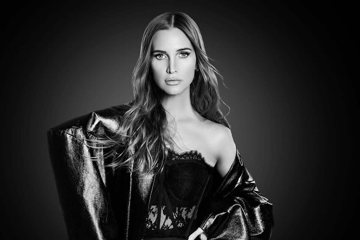 Ashlee Simpson in black leather jacket and lace top standing in front of black background looking into the camera. 