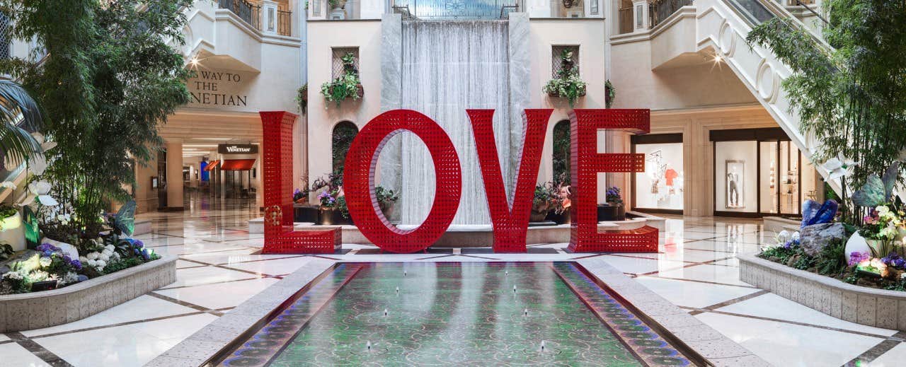 A large "LOVE" sculpture stands in the center of an elegant mall with shops, greenery, and a decorative water feature.