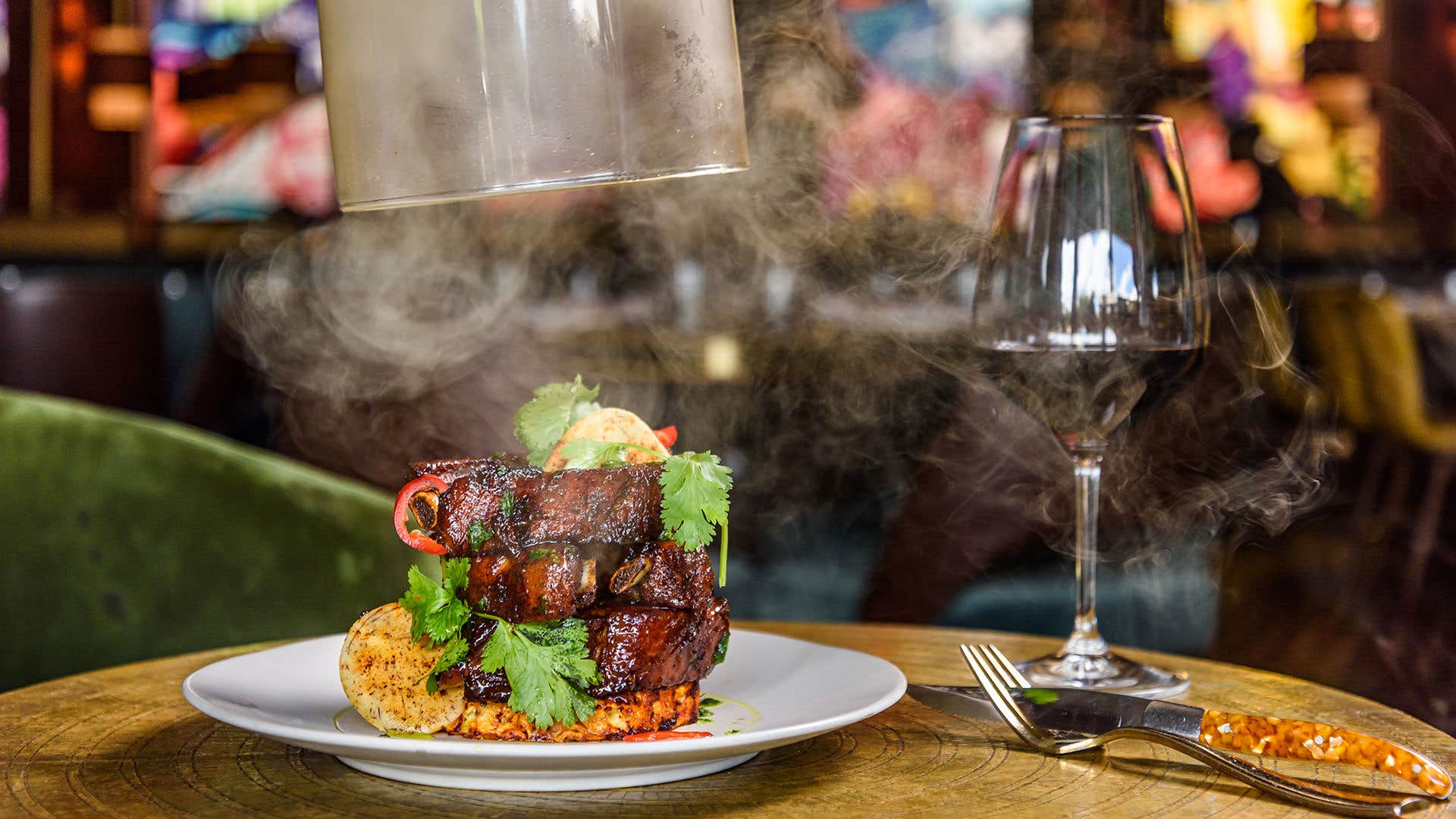 Smoke escapes as a glass dome uncovers a baby back ribs dish on a table with a glass of wine.