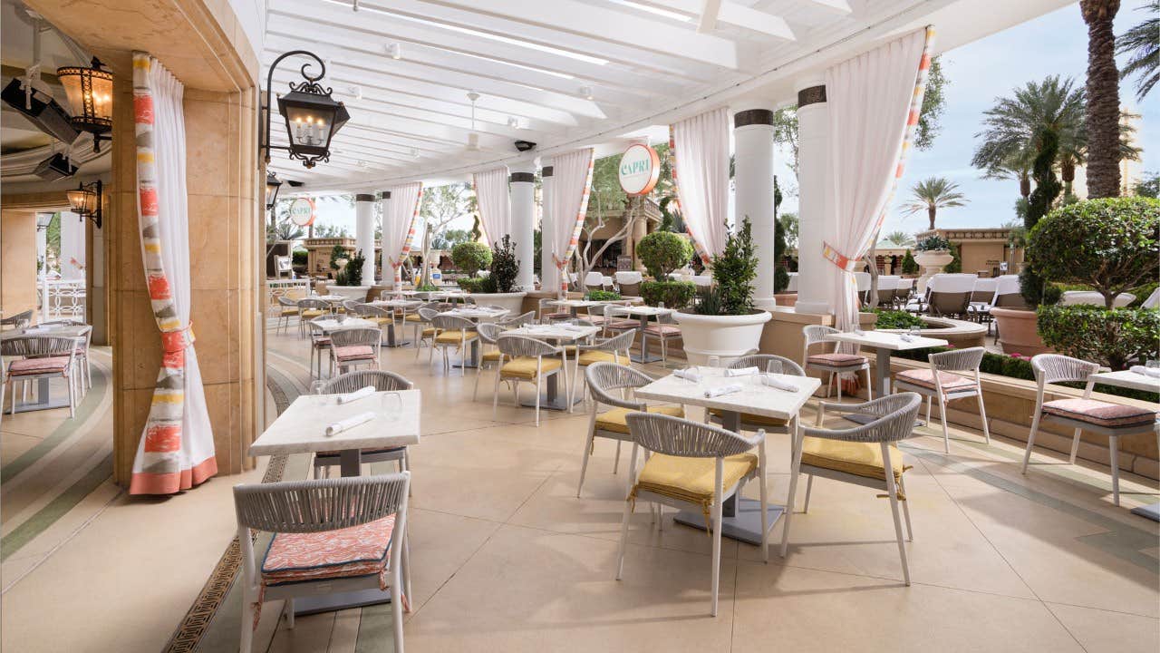 Outdoor restaurant patio with elegantly set tables, chairs, and white curtains, surrounded by greenery and palm trees.