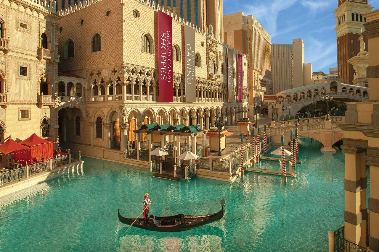 A gondola on a canal near a Venetian-style building with shops and restaurants, adorned with striped poles and banners.