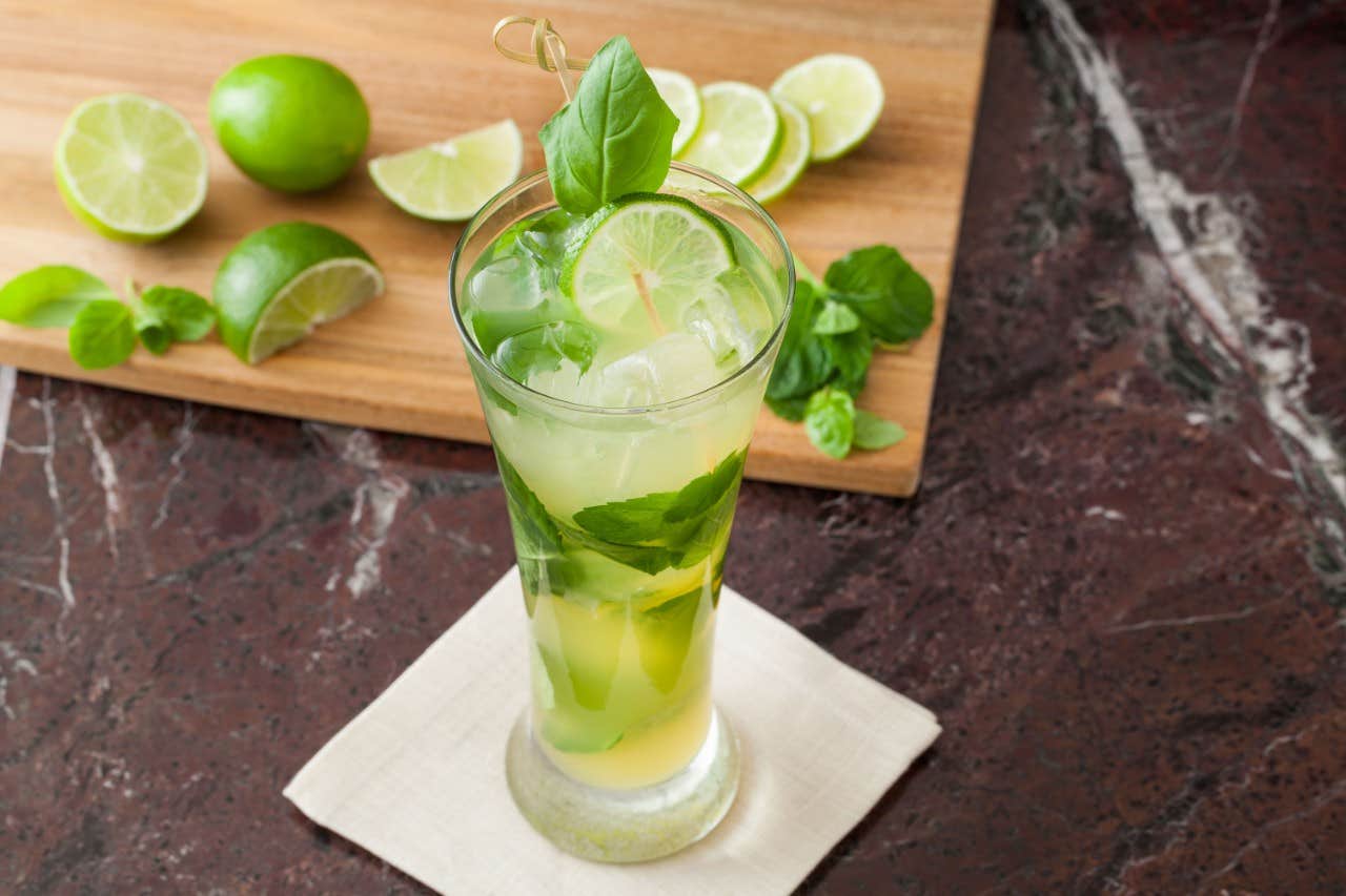 Tall glass of mojito with lime slices and mint, placed on a napkin, with a cutting board, lime, and mint in the background.