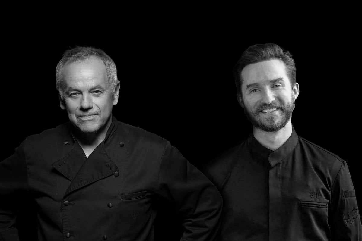 Two people wearing dark chef jackets standing side by side against a black background.