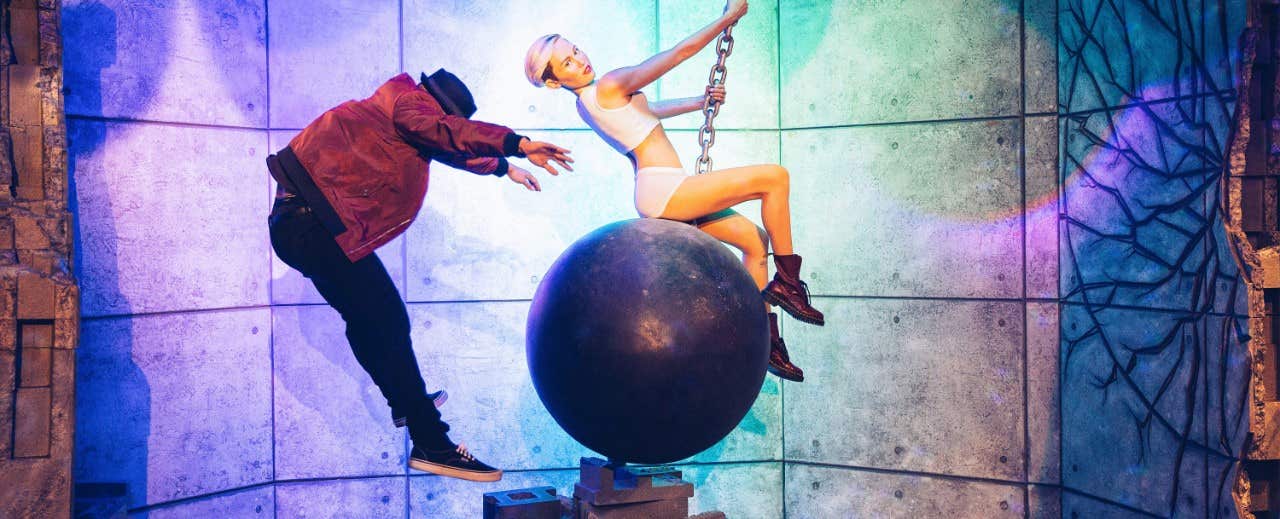 A person in a dark jacket leans back dramatically while another in white sits and hangs on to a large wrecking ball.