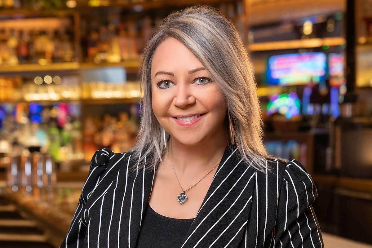 Woman with gray hair in a black and white striped jacket smiles in a bar setting with blurred background.