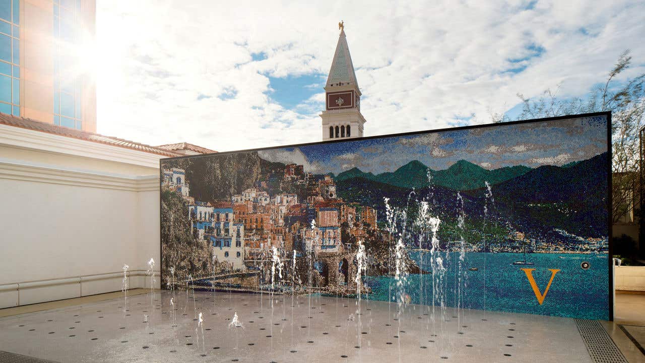 Outdoor fountain plaza with small vertical water jets in front of a large mural depicting a coastal town, with a tall tower visible behind the wall.