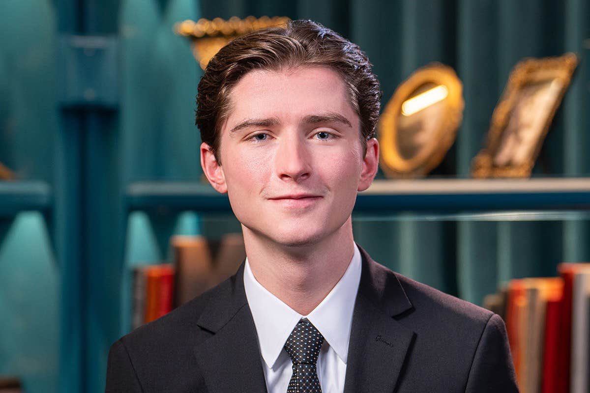 A young man in a suit and tie stands in front of a bookshelf filled with framed pictures and books.