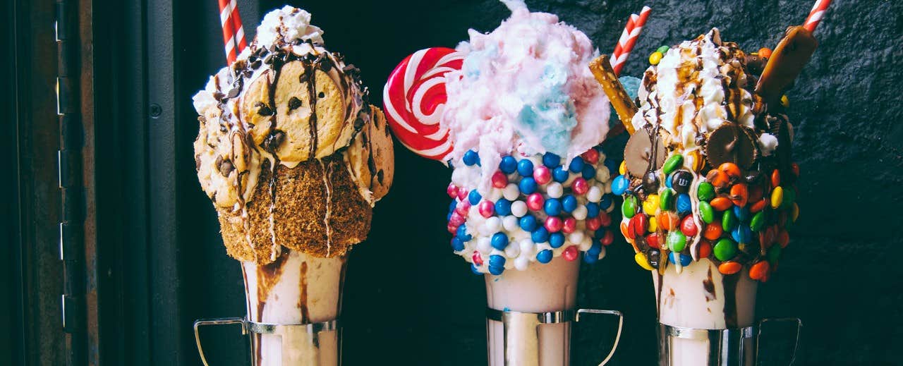 Three extravagant milkshakes topped with cookies, candy, and cotton candy, each garnished with whipped cream and drizzles.