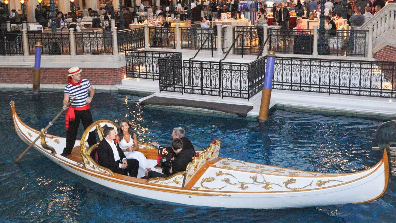 Four people dressed formally in a gondola driven by a gondolier on a blue canal, with a busy balcony in the background.