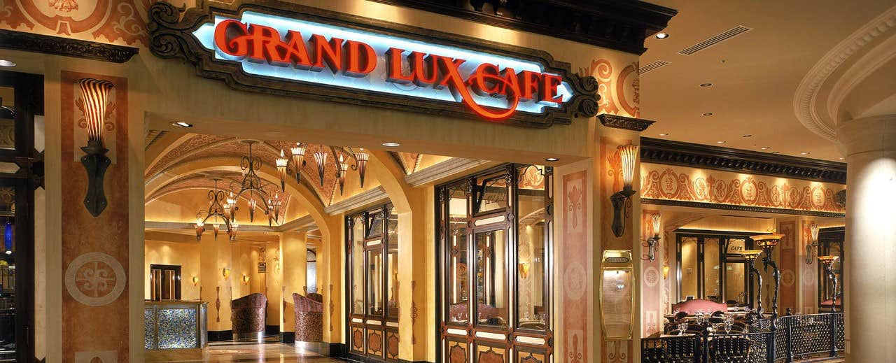 Entrance of the Grand Lux Cafe with elegant decor, chandeliers, and a welcoming illuminated sign above the doorway.