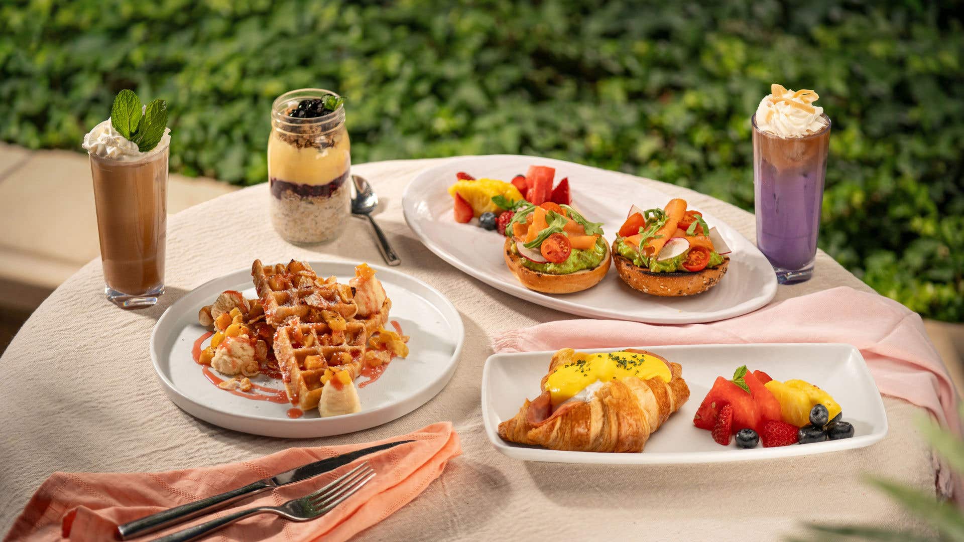Assorted brunch dishes on an outdoor table, including waffles, avocado toast, a croissant with eggs, fresh fruit, and an iced coffee.