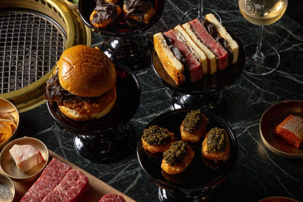 A luxurious meal spread on a dark marble table, featuring a burger, sandwiches, caviar, and raw marbled beef next to a tabletop grill.