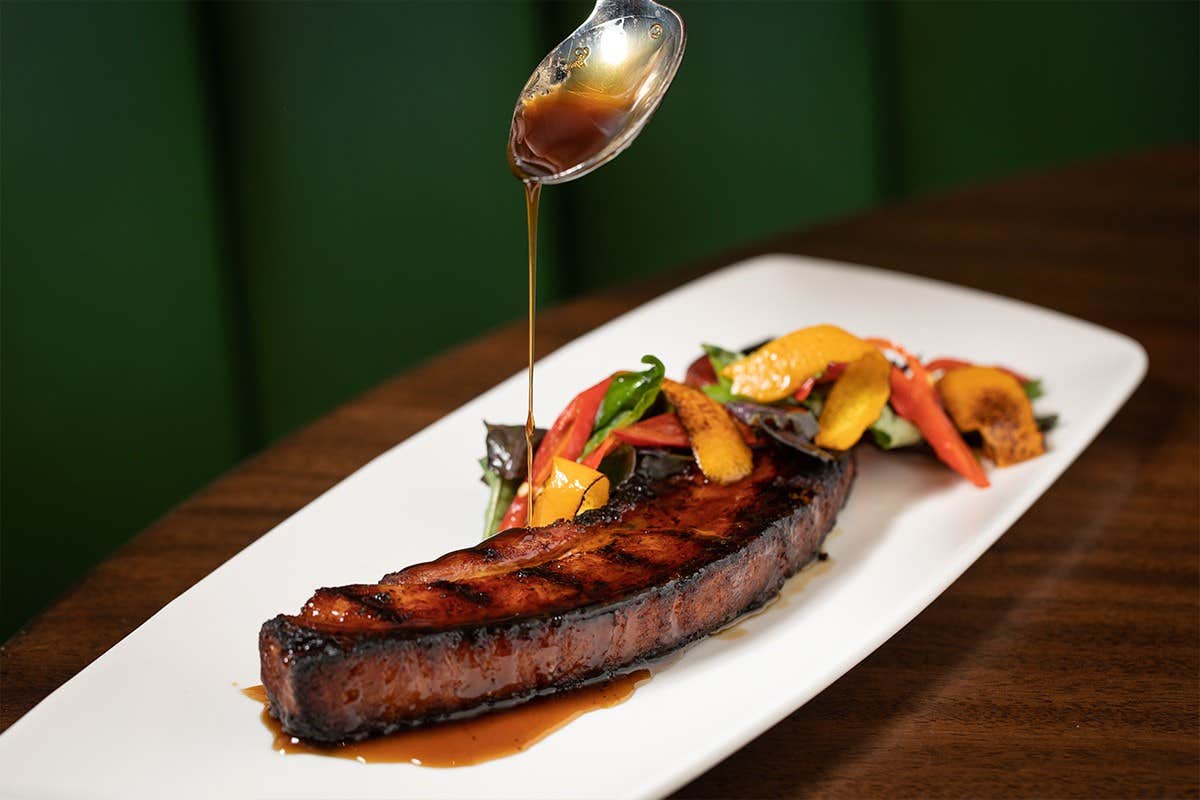 A grilled meat slice with vegetables on a white plate being drizzled with sauce from a spoon.