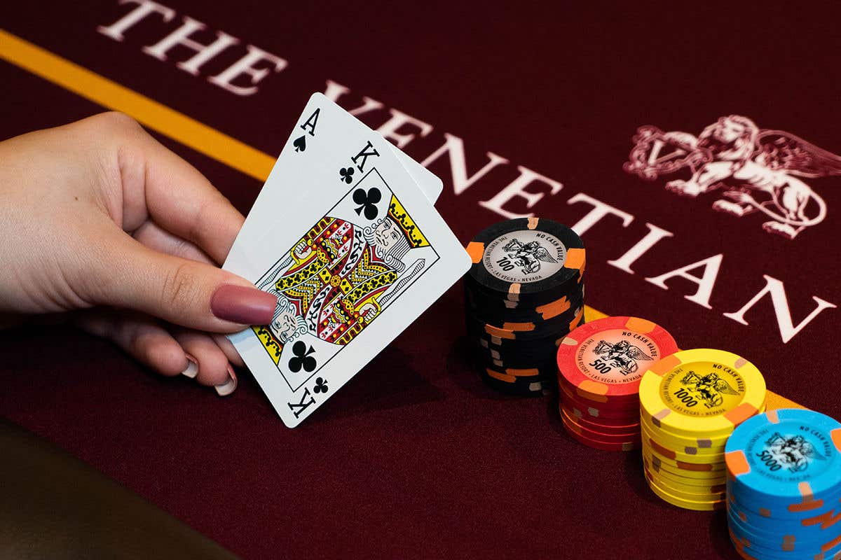 A hand holds an ace and king of clubs beside stacks of colorful casino chips on a red Venetian gaming table.