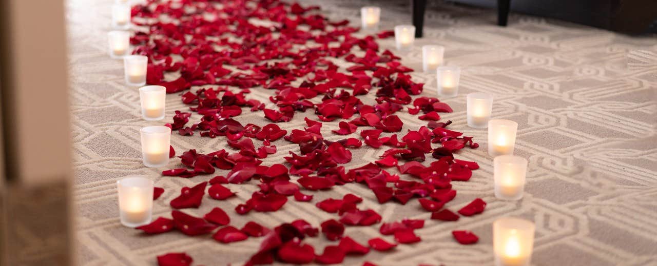 A carpeted walkway covered with scattered red rose petals and lined with lit white candles in glass holders.