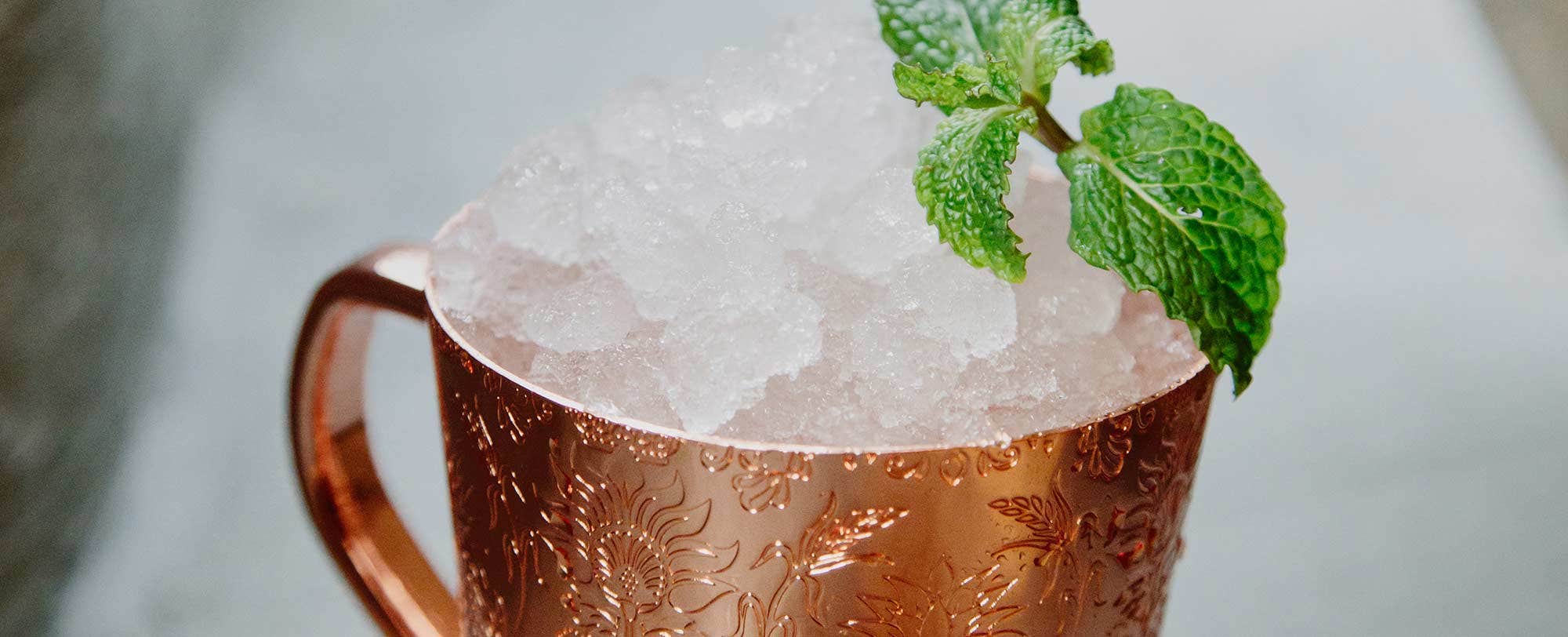 Copper mug with crushed ice and a sprig of mint on top, details of floral engravings visible on the mug.