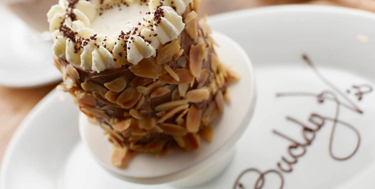 A tall dessert covered in almond slices and topped with whipped cream on a white plate with cursive writing.