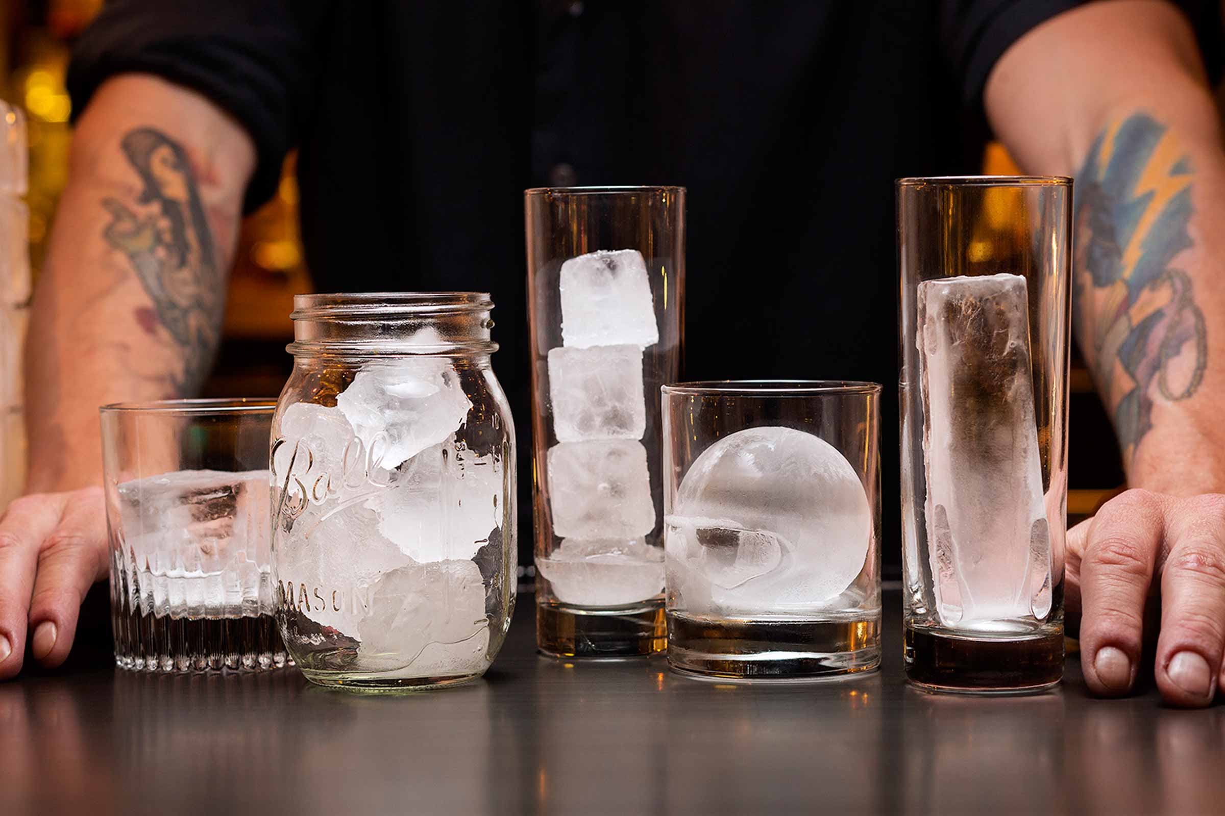 Various glasses with different shapes of ice on a counter. A person with tattooed arms is in the background.