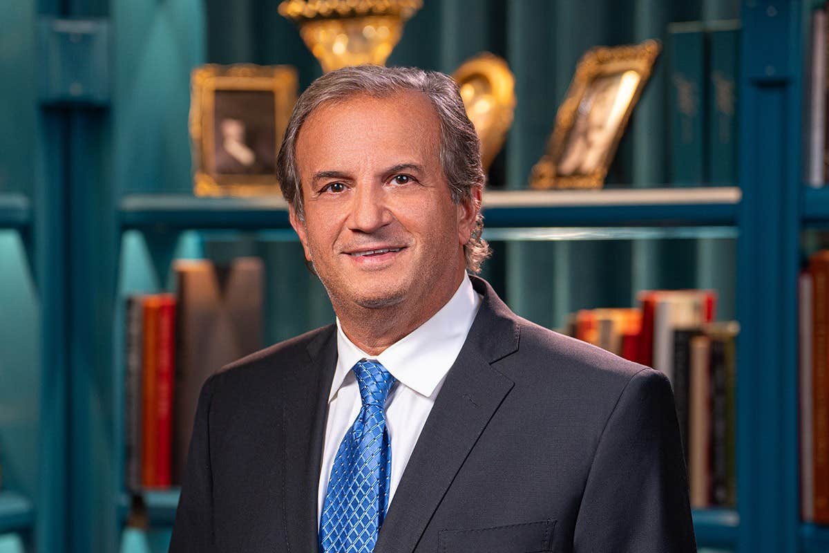 Man in a suit with a blue tie standing in front of a bookshelf with framed photos and books.